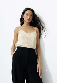 Débardeur en soie beige avec dentelle et décolleté en V, associé à un pantalon noir taille haute. Comprend des bretelles fines et une texture lisse.