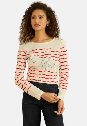 Světle béžový svetr s dlouhým rukávem s červenými vlnitými pruhy, s vyšitým nápisem „la Mer“ v modré barvě a zlatými hvězdnými akcenty poblíž ramene.