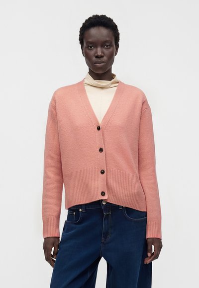 Femme portant un cardigan rose clair boutonné par-dessus un col roulé crème et un jean bleu foncé sur fond blanc uni.