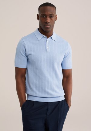 Muž v světle modré krátkosleevé pletené polokošili a tmavě modrých kalhotách stojící s rukama v kapsách na neutrálním pozadí.