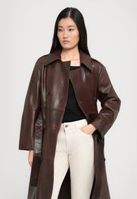 Femme avec de longs cheveux noirs portant un trench-coat en cuir marron foncé sur un haut noir et un pantalon cream taille haute, avec une main dans la poche.