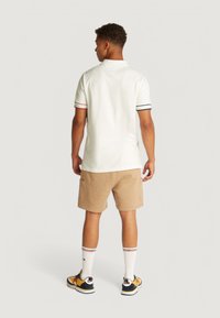 Camisa polo blanca con acentos a rayas rojas y marinas, combinada con pantalones cortos beige. El modelo está de pie de espaldas, mostrando calzado casual y calcetines.