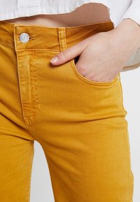Jeans de mezclilla amarillos mostaza con corte ajustado, diseño de cinco bolsillos, costuras visibles y cierre de botón plateado en la cintura.