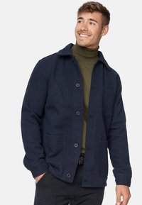 Mørkeblå jakke lavet af tekstureret stof med krave, knaplukning og to forlommer, lagdelt over en olivengrøn turtleneck.