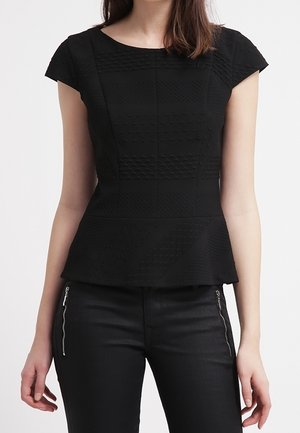 Frau trägt ein eng anliegendes schwarzes, strukturiertes Kurzarmtop mit Taillen-Peperlume, kombiniert mit schwarzen Hosen mit Reißverschlussdetails vorne.