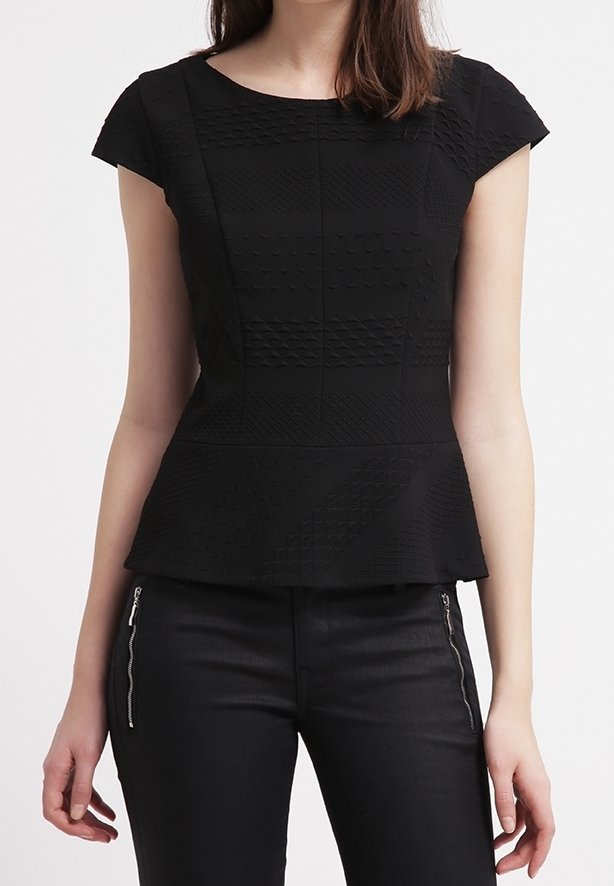 Kobieta nosi dopasowaną czarną teksturowaną bluzkę z krótkim rękawem i baskinką w talii, zestawioną z czarnymi spodniami z ozdobnymi suwakami z przodu.