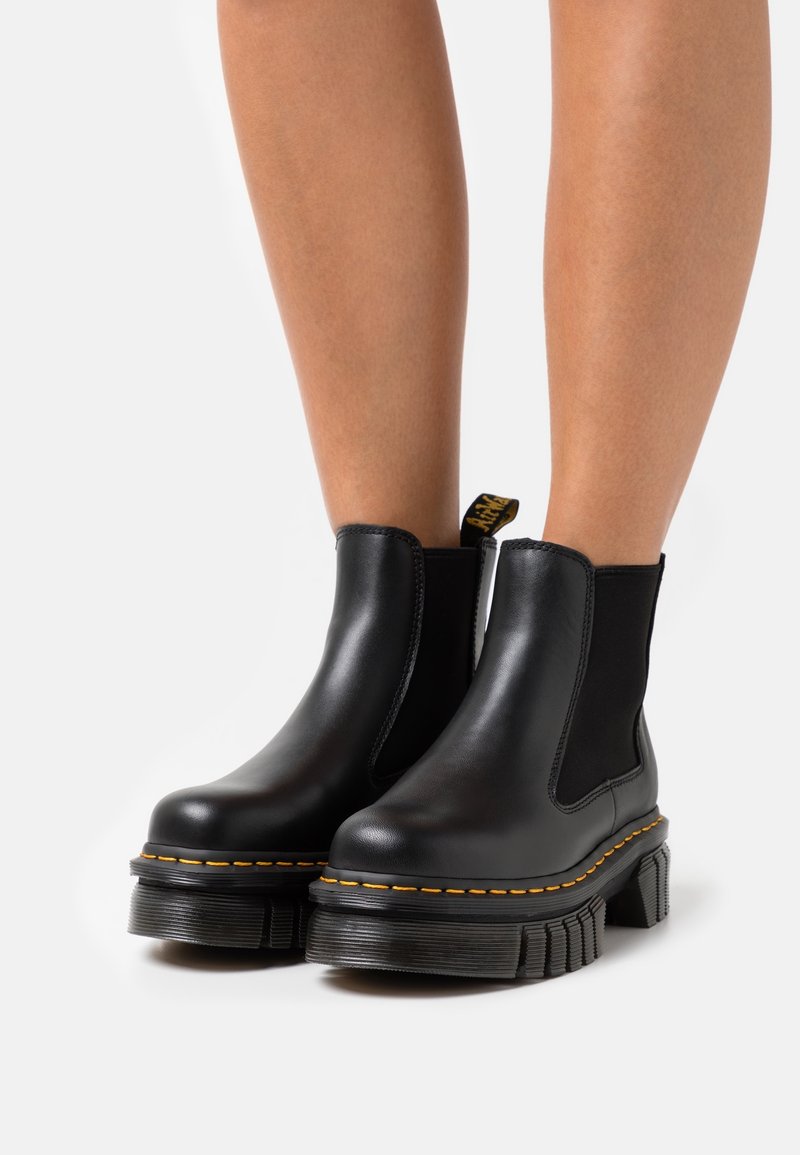 Bottines en cuir noir avec semelles plateformes épaisses et rainurées, coutures jaunes, portées sur des jambes nues sur fond blanc.