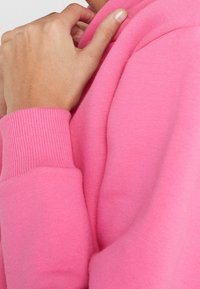 Sudadera rosa ajustada en tela suave con puños acanalados. Primer plano muestra una mano apoyada en el escote, destacando la textura y el color.