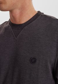 Mørkegrå sweatshirt med ribbet krave og et diskret V-hals design. Har en cirkulær sort logo-patch på venstre bryst. Blød tekstur.