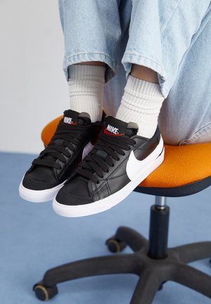 Černé kožené tenisky s bílou podrážkou a swoosh, černé tkaničky a červené logo na jazyku, spárované s bílými žebrovanými ponožkami a modrými džíny.