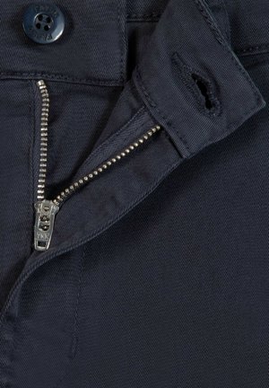 Close-up van marineblauwe stof met een gedeeltelijk open zilveren rits en een zwarte knoop met het label "FAGUA" op een tailleband.