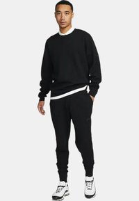 Completo felpa nera e pantaloni della tuta, con dettaglio del collo interno bianco, polsini elasticizzati e accenti con logo. Indossato con sneakers bianche.