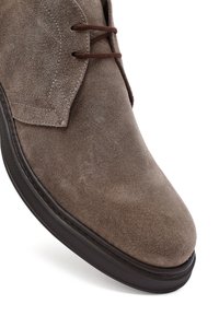 Zapato de hombre lace-up de ante marrón con suela de goma negra, primer plano en la punta y área de cordones sobre un fondo blanco.