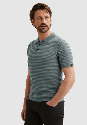 Mann mit Bart, der ein hellblaues strukturiertes kurzärmeliges Poloshirt und schwarze Hose trägt, die Hand in der Tasche, schaut zur Seite.