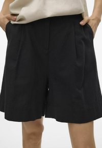 Pantaloni corti neri in lino con pince frontali, tasche laterali e una vestibilità ampia. Il tessuto presenta una texture sottile e un design pulito e classico.