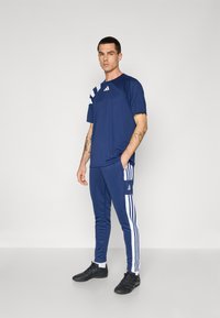 Camiseta atlética marina con franjas laterales blancas y logotipo, combinada con pantalones marinos con franjas y logotipo blancos. Zapatillas deportivas negras.