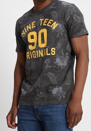 Hombre con camiseta gris oscuro con estampado floral y texto amarillo "NINE TEEN 90 ORIGINALS" y vaqueros azules.