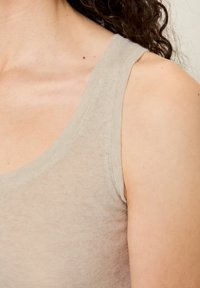 Šviesiai smėlio spalvos liemenėlė su apvaliu iškirpimu ir siaurais petnešėlėmis, pagaminta iš minkštos, tekstūruotos medžiagos. Siūlių detalės matomos palei kraštus.