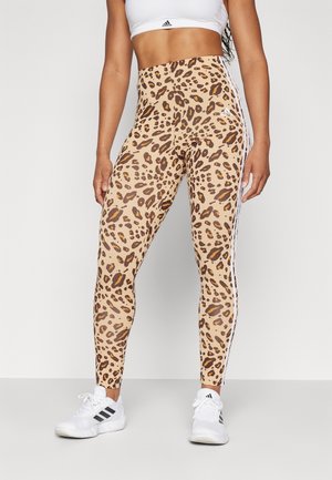 Leggings taille haute en imprimé léopard beige avec des taches brunes, dotés de bandes latérales blanches et portés avec des chaussures de sport blanches.