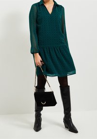 Teal patterned dress with a V-neck, long sheer sleeves, and a gathered skirt. Paired with black knee-high boots and a black handbag.