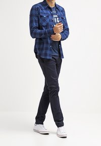 Modrá kostkovaná flanelová košile s knoflíkovými kapsami, nošená přes námořnické tričko, v kombinaci s tmavými kalhotami a bílými teniskami.