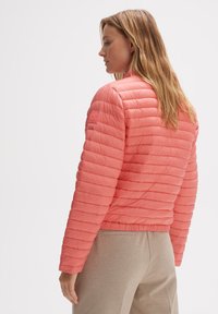 Veste matelassée en corail avec un design à quilting horizontal, col montant et ourlet élastique. Le matériau semble lisse et léger, adapté pour le superposition.