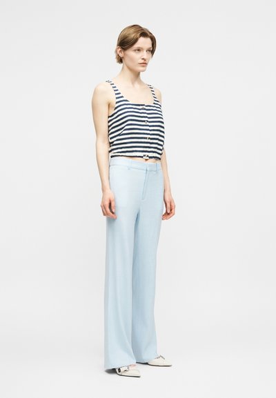 Γυναίκα που φοράει μπλε-ναυτικό και λευκό ριγέ αμάνικο κοντό τοπ, ανοιχτό μπλε φαρδιά παντελόνια και λευκά μυτερά παπούτσια, στέκεται μπροστά σε μια απλή φόντο.