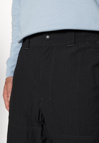 Homme portant un pantalon noir avec un bouton pression et des passants de ceinture, associé à une chemise bleu clair à manches longues, main reposant sur le côté.