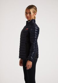 Marineblaue Daunenjacke mit hohem Kragen, schlanker horizontal gestepptem Design und einem Logo-Patch auf der Brust. Glatte, glänzende Textur.
