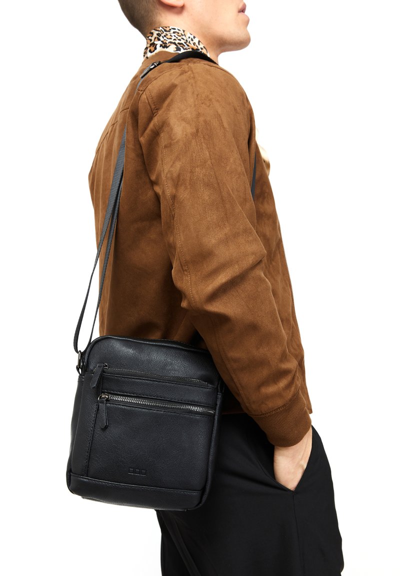 Borsa a tracolla in pelle nera con due tasche frontali con zip, texture liscia, forma rettangolare compatta, indossata sulla spalla da un modello.