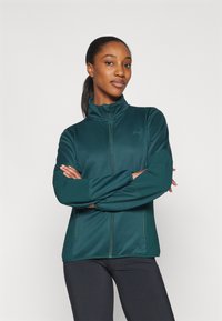 Giacca zip-up di colore teal realizzata in tessuto morbido ed elastico. Presenta un colletto alto e maniche lunghe. Il modello indossa leggings neri.