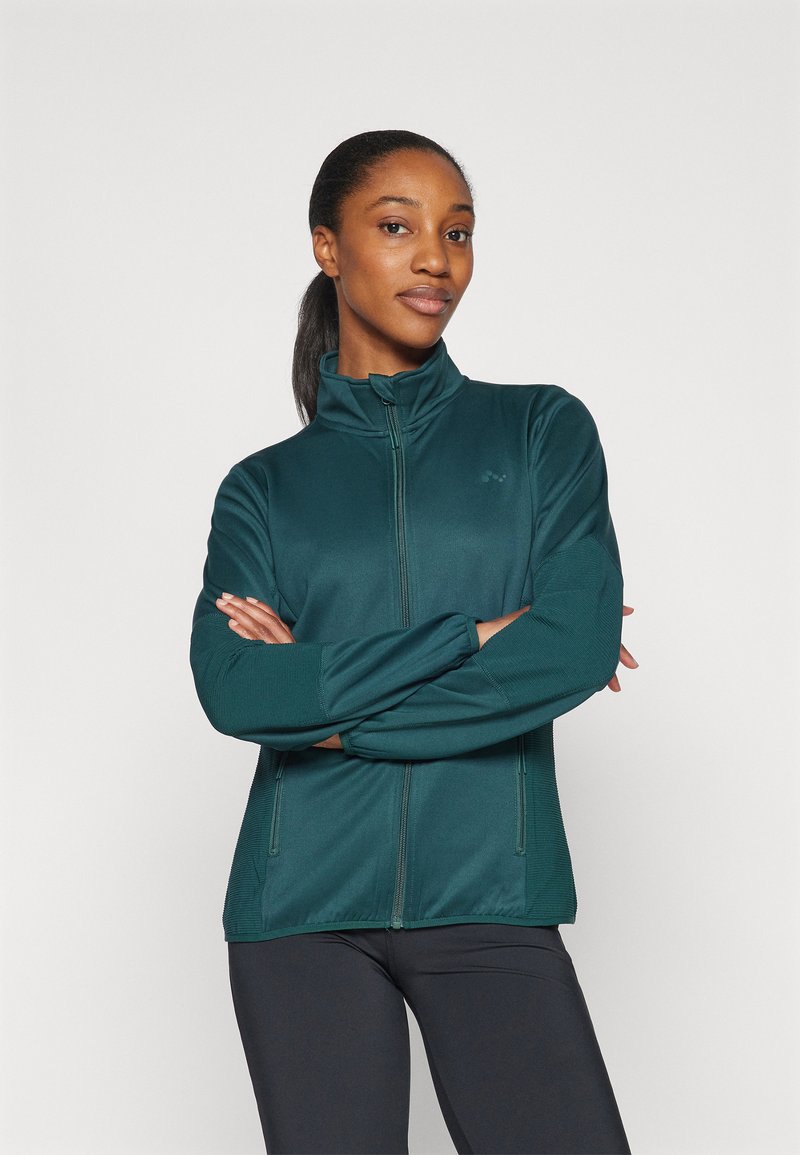 Giacca zip-up di colore teal realizzata in tessuto morbido ed elastico. Presenta un colletto alto e maniche lunghe. Il modello indossa leggings neri.