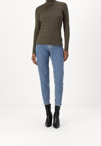 Personne portant un pull côtelé à col roulé vert olive, un jean droit bleu et des bottines noires à talons pointus se tenant sur un fond blanc.