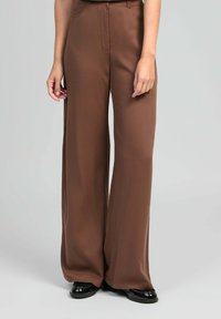 Pantalons marron à jambes larges en tissu lisse, avec taille haute, plis avant et fermeture éclair. Complétés par des chaussures noires.