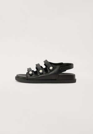 Black leather sandal featuring three adjustable straps with metal accents, flat sole, and an open back design. Smooth texture overall.