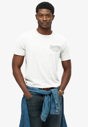 Camiseta gris de manga corta con un logo impreso en el lado izquierdo del pecho, combinada con jeans oscuros y una camisa de mezclilla azul clara atada a la cintura.