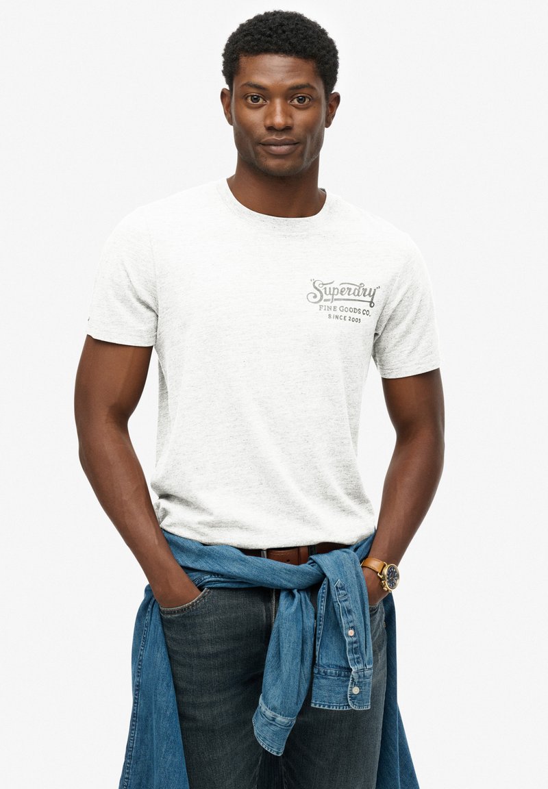 Camiseta gris de manga corta con un logo impreso en el lado izquierdo del pecho, combinada con jeans oscuros y una camisa de mezclilla azul clara atada a la cintura.