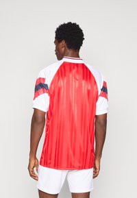 Camisa de futebol vermelha com riscas verticais, mangas curtas brancas com detalhes em preto e vermelho, e um colarinho em contraste. Tecido liso e brilhante.