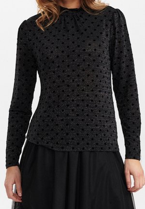 Žena v přiléhavém černém tričku s dlouhým rukávem s vystouplými puntíky a jemným leskem, doplněném černou sukní.