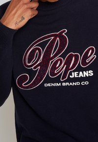 Maglione a maglia blu navy con un grande logo "Pepe" rosso glitterato e testo bianco "DENIM BRAND CO", con polsini e scollatura a costine.