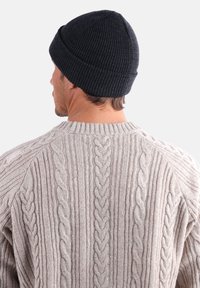Homme portant un bonnet en tricot noir et un pull en tricot torsadé beige, vu de dos contre un fond clair uni.