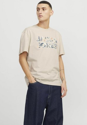 Beige bomulds t-shirt med korte ærmer, der har et flerfarvet grafisk print inklusive teksten "JACK & JONES." Båret med mørke denimbukser.