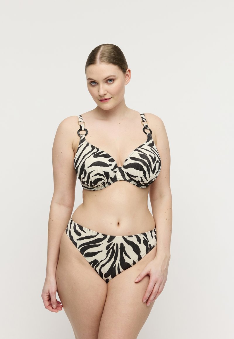 Frau mit heller Haut und Haaren in einem Bikini mit Zebra-Muster, die vor einem einfarbigen weißen Hintergrund steht und in die Kamera blickt.