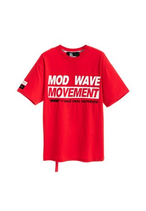 Joven con cabello claro que lleva una camiseta roja con el texto "MOD WAVE MOVEMENT", pantalones anchos blancos y zapatos marrones, posando frente a un fondo blanco liso.