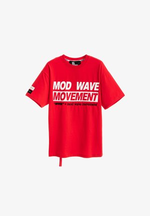 Joven con cabello claro que lleva una camiseta roja con el texto "MOD WAVE MOVEMENT", pantalones anchos blancos y zapatos marrones, posando frente a un fondo blanco liso.