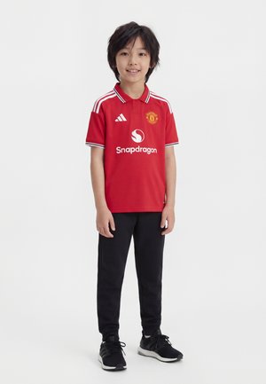 Jeune garçon debout, portant un maillot de football rouge de Manchester United avec des logos, un pantalon noir et des baskets noires, souriant à la caméra.