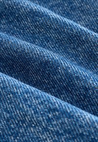 Denim stof in blauwe tinten, met een textuur waarbij de stiknaden zichtbaar zijn en subtiele kleurvariaties in de weving aanwezig zijn.