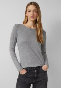 Young woman with long blonde hair wearing a gray long-sleeve top and black jeans, standing with hands in pockets against a plain background.
