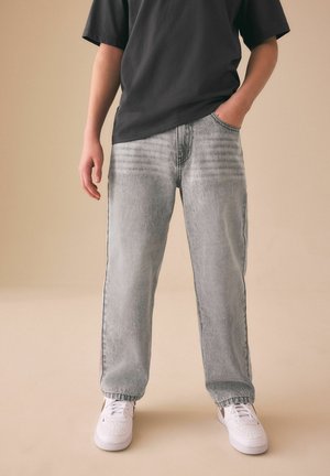 Person iført lysegrå vasket jeans, hvide sneakers og en sort kortærmet skjorte, med den ene hånd i lommen, stående på en beige gulv.
