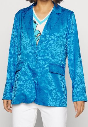 Vrouw draagt een felblauwe jacquardblazer met bloemenprint over een gebreide V-hals trui met patroon en witte broek, getoond van schouders tot heupen.